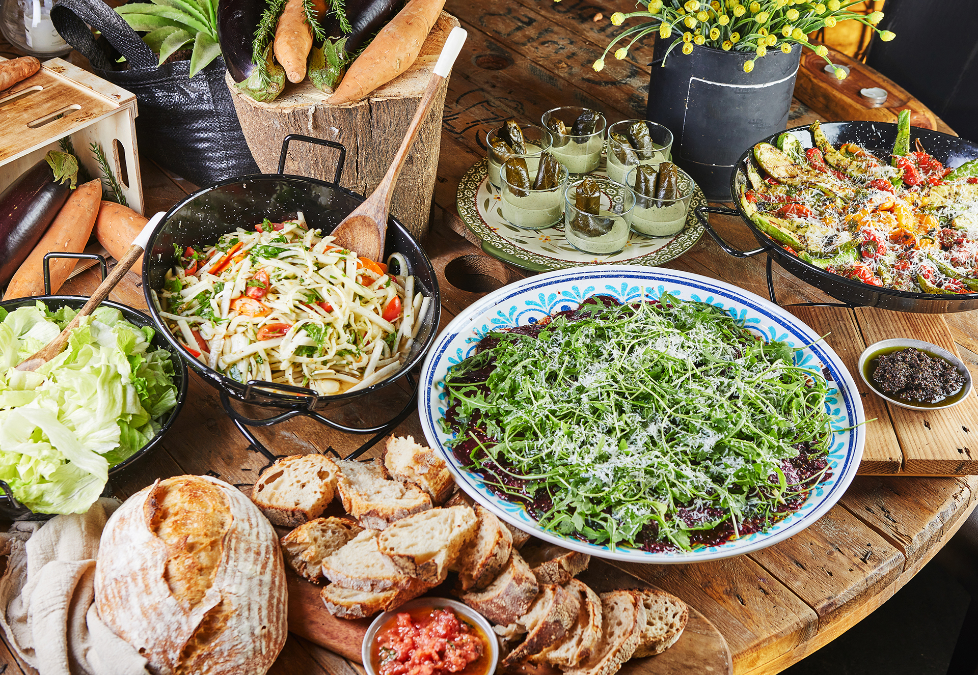 Rustikaler Holztisch mit verschiedenen Gerichten, darunter frischer Salat, gemischtes Gemüse, Brot und gefüllte Weinblätter in Gläsern, dekoriert mit geriebenem Käse und Kräutern, umgeben von Süßkartoffeln und anderen frischen Zutaten.