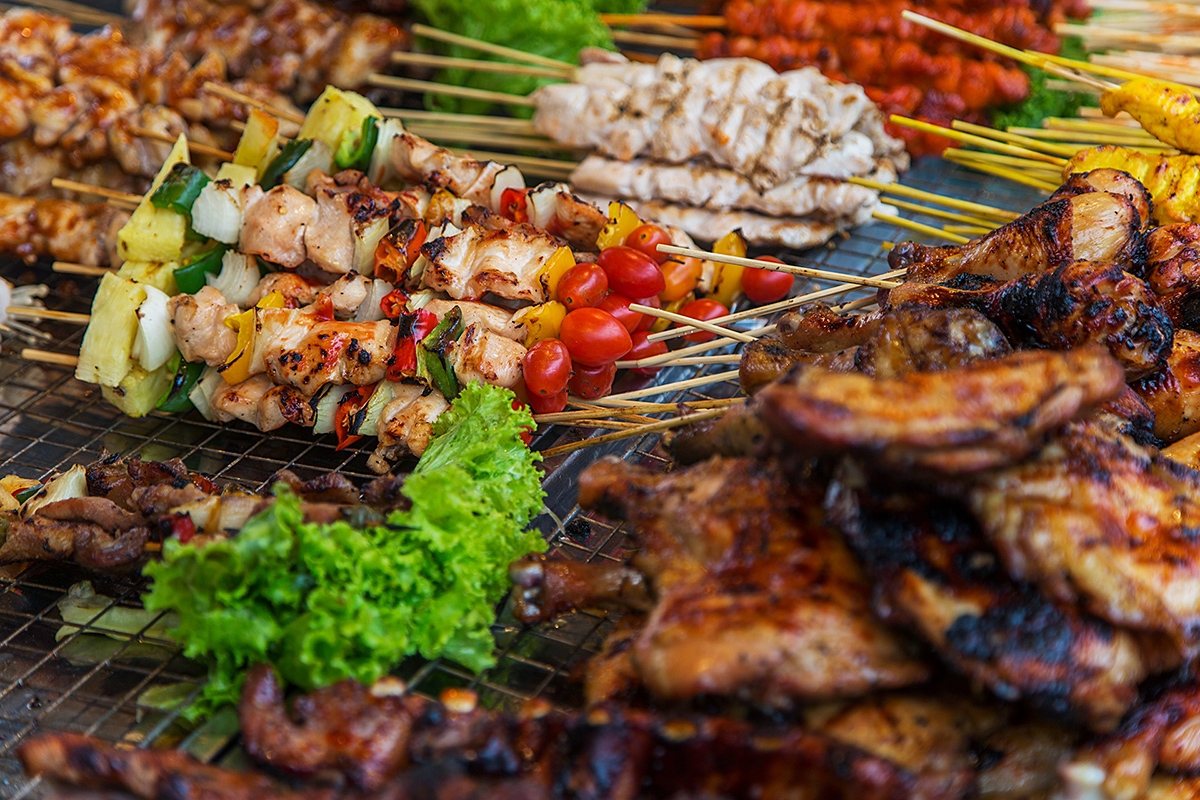 Verschiedene gegrillte Fleischspieße mit Gemüse und frischem Salat auf einem Grillrost, darunter Hühnchenspieße mit Paprika, Tomaten und Ananas sowie Hähnchenflügel.